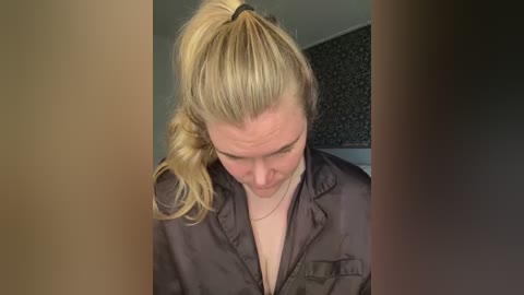 A video of a blonde woman with a ponytail, wearing a black shirt, looking down with a slight smile. Background shows a dark patterned wall and a white door.
