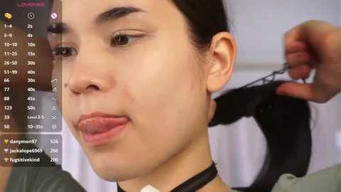 Video of a young woman with light skin, dark hair, and a black choker, having her hair pulled by a hand holding a comb. Background shows a bathroom with a white wall and pink and purple icons on a screen.