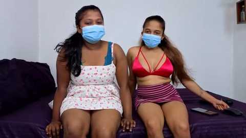 Video of two young women, one in a floral dress, the other in a red bikini top and striped skirt, sitting on a bed in a plain bedroom.