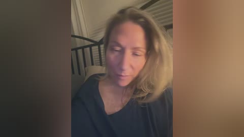 Video of a middle-aged Caucasian woman with blonde hair, wearing a dark top, standing in a dimly lit room with a black metal bed frame and closed blinds in the background.