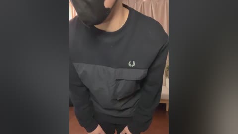 Video of a person wearing a black long-sleeve sweatshirt with a black mask, standing in a dimly lit room with a white curtain and a wooden floor.