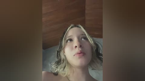 Video of a blonde woman with wavy hair, fair skin, and closed eyes, lying on her back in a dimly lit room with wooden paneling and a bedsheet.