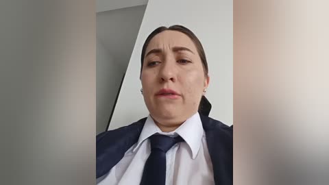 Video of a middle-aged, light-skinned woman with short brown hair, wearing a white shirt and dark tie, sitting in a dimly lit room with blurred walls, giving a serious expression.