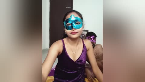 Media: Video of a young woman with light skin, dark hair, and purple dress, wearing a blue and white masquerade mask. Behind her, a man with a similar mask and another person are partially visible.