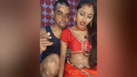 Video of a young Indian couple in traditional attire, the woman wearing a red sari with intricate gold embroidery and jewelry, and the man in a blue shirt, both posing intimately.