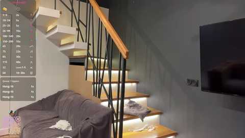 A video of a modern, minimalist living room with a wooden staircase, grey sofa, wall-mounted TV, and a gray throw blanket.