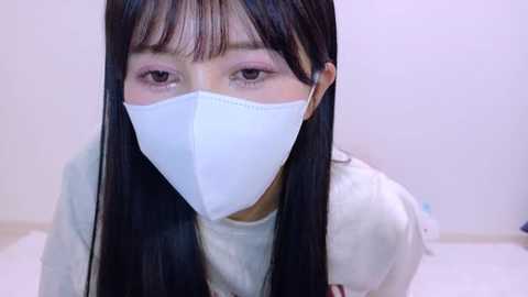 Video of an East Asian woman with long black hair, wearing a white surgical mask, gray sweater, and pale pink eyeshadow, in a sterile white room with minimal background details.