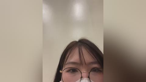 A video of an Asian woman with straight black hair, wearing round glasses, and a face mask, partially visible, against a blurred beige background.
