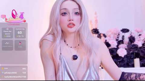 Video of a young woman with platinum blonde hair, wearing a revealing silver halter top, standing in a room adorned with pink and black flowers.
