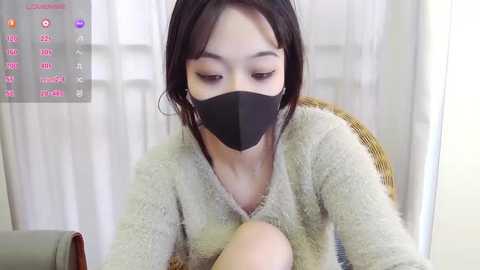 A video of an Asian woman with fair skin, short black hair, and a black face mask, wearing a beige fuzzy sweater. She is sitting indoors with white curtains in the background.
