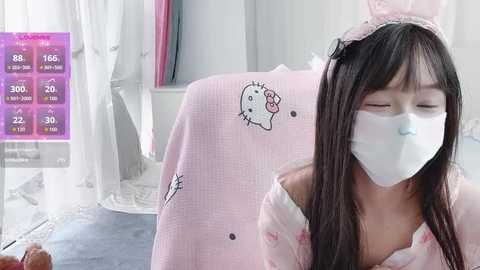 Video of an East Asian woman in a pink Hello Kitty mask and robe, with long dark hair, eyes closed, sitting indoors, background featuring soft lighting and a digital screen displaying health data.