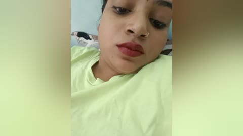 Media: A close-up video of a young woman with medium brown skin, wearing a light green t-shirt, lying on a bed. Her eyes are partially closed, and she has a subtle nose ring. The background shows a soft, light blue wall.
