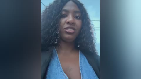Video of a young Black woman with curly hair, wearing a light blue top, standing in a dimly lit hallway, with blurred background.