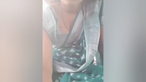 Media: Video of a woman wearing a teal saree with white polka dots, draped over her left shoulder, and a light blue top. Her face is partially visible, and she has a dark complexion. The background is out of focus.