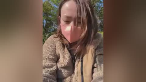 Video of a young woman with fair skin and shoulder-length brown hair, wearing a pink face mask, a beige jacket, and a tan vest, outdoors with blurred green foliage in the background.