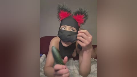 Video of a person in a black mask with red cat ears, holding a cucumber, on a bed with a striped blanket.