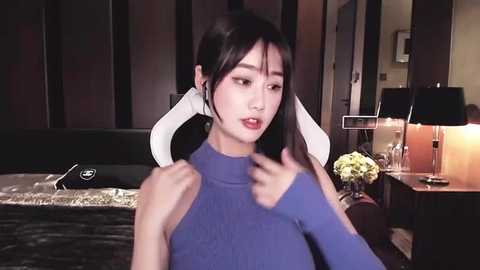 Video of a young Asian woman with straight black hair, wearing a blue turtleneck sweater, in a dimly lit room with dark furniture, a bed, and a lamp.