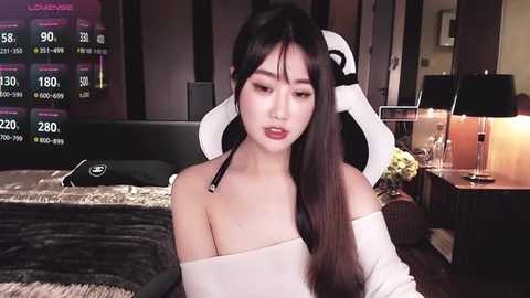 Video of a young East Asian woman with long, straight black hair and fair skin, wearing a white off-shoulder top. She sits in a modern bedroom with a black headboard, a digital screen displaying stock market data, and a white gaming chair.