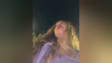 Video of a blonde woman with wavy hair, wearing a light-colored top, gazing upwards with a serene expression. The background is a dark, blurred night sky.