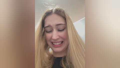 Media: Video of a young Caucasian woman with long, blonde hair, smiling with closed eyes, taken from a slightly above angle, showing her from the shoulders up against a blurred background.