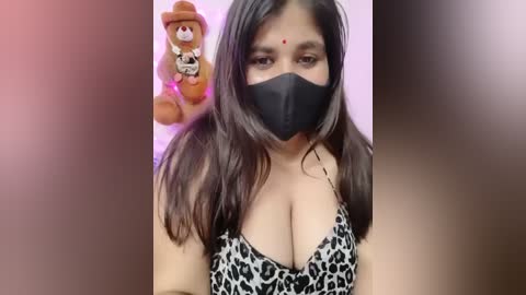 Media: Video of a South Asian woman with long dark hair, wearing a black face mask and leopard-print top, with a stuffed bear in the background.