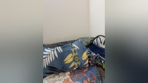 A video of a corner of a bed with colorful, patterned pillows and blankets in blue, green, and orange hues against a plain white wall.