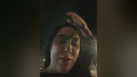 A dimly lit, grainy video of a woman with long dark hair, wearing a black sweater, sitting with her hand on her forehead, giving a slight smile. The background is dark and blurry.