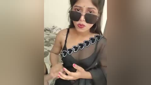 Media: Video of a young South Asian woman with fair skin, wearing black sunglasses, a sheer black saree with silver embroidery, and red lipstick, standing indoors with a blurred background.