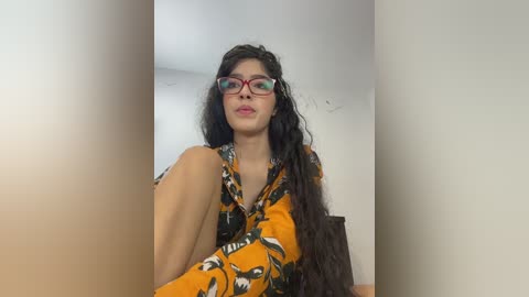 Video of a young woman with long, wavy black hair and fair skin, wearing red-rimmed glasses and a mustard-yellow shirt with a black wolf pattern. She sits on a dark wooden chair against a plain white wall, with a subtle out-of-focus background.