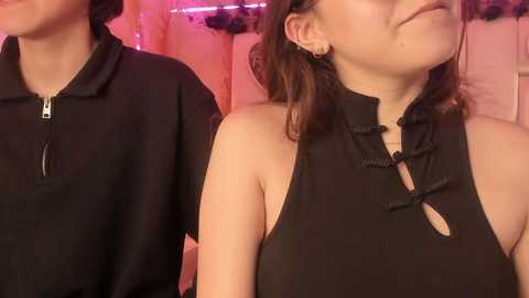 Video of two women wearing black, sleeveless, high-neck dresses with intricate, decorative bows. One woman has short, dark hair, while the other has shoulder-length, wavy hair.