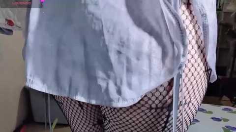 A video of a person wearing a light blue button-up shirt, fishnet stockings, and a white bra. The image is taken indoors, with a blurred background featuring kitchen appliances.