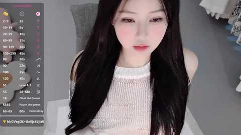 Video of an Asian woman with long black hair, fair skin, and large eyes wearing a white knit top, captured on a phone screen with a live stream overlay.