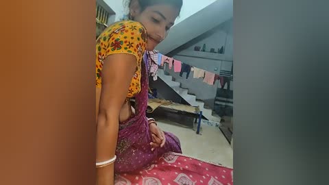 Video of a South Asian woman in traditional attire, wearing a colorful sari and a short-sleeved top, sitting in a cluttered, dimly lit room with a staircase and laundry hanging.