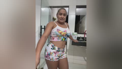 Video of a young woman with light brown skin, wearing a colorful butterfly-patterned crop top and shorts, standing in a modern, tiled bathroom.