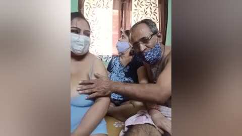 Media: Video of a man and a woman in a hospital room, both wearing masks and light blue gowns. The man, with glasses and gray hair, cradles the woman's breast, while she gazes at him with concern.
