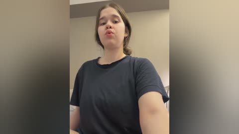 Video of a young woman with light skin and brown hair, wearing a dark blue t-shirt, making a pouty face, indoors against a beige wall.