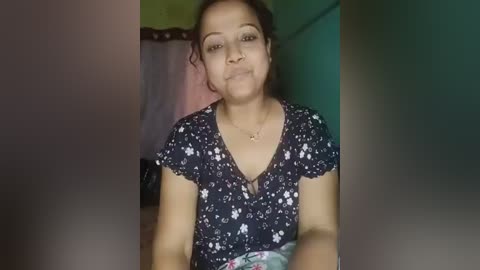 Video of a middle-aged South Asian woman with medium-brown skin, wearing a floral-patterned dark blouse, seated indoors. Background features a green wall and a pink curtain.