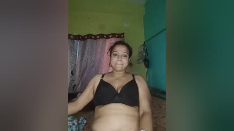 A video of a pregnant woman in a black bra, sitting on a bed with a teal wall and a yellow-green wall. The room has a pink and white curtain, and a patterned bedspread.