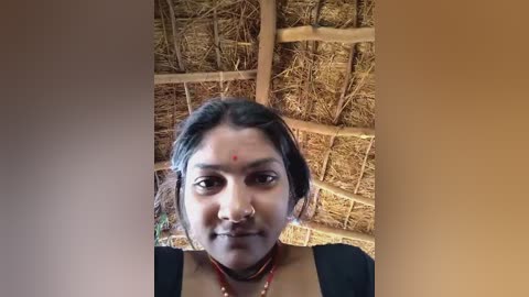 Media: A video of a South Asian woman with medium brown skin, black hair, and a nose ring, wearing a black top, standing in a rustic, thatched-roof hut.