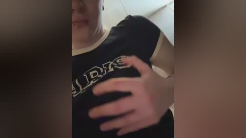 Video of a woman with fair skin wearing a black t-shirt with white lettering, holding her left breast. The background is blurred, with an indistinct beige wall.