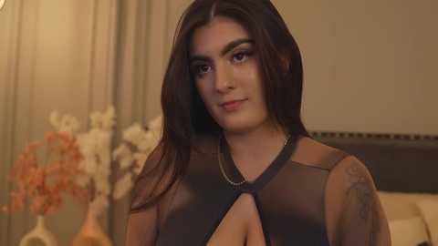 Video of a young woman with long, dark hair, medium skin tone, wearing a sheer, black, plunging neckline top, standing indoors, with blurred floral arrangements in the background.