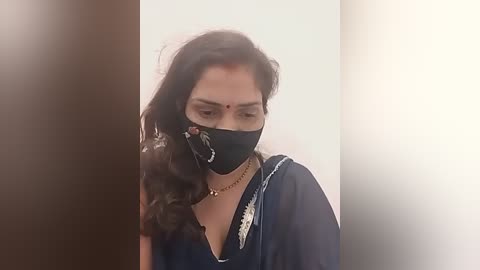 Media: A video shows a woman with long, wavy black hair, wearing a black face mask, a black top with white floral embroidery, and a gold necklace, indoors with a plain white background.
