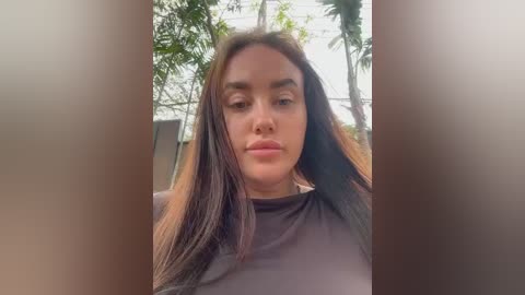 Media: A video of a woman with long brown hair, light skin, and brown eyes, wearing a black top, taken outdoors with blurred greenery in the background.