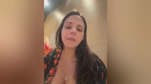 Video of a smiling, light-skinned woman with long dark hair, wearing a floral robe, standing in a dimly lit room with beige walls.