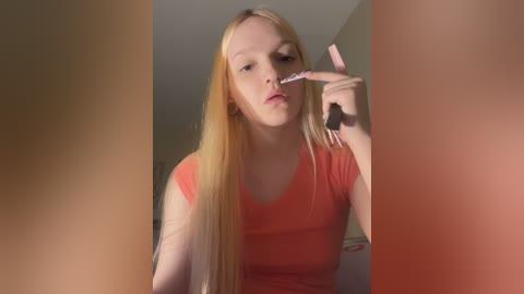 Video of a young blonde woman with pale skin, wearing a red T-shirt, brushing her teeth with a pink toothbrush in a bathroom with beige walls.