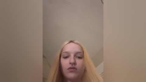 Video of a young Caucasian woman with long, straight, platinum blonde hair, wearing minimal makeup, standing in a dimly lit room with beige walls and a circular ceiling light.