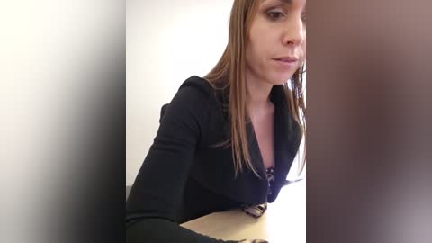 Video of a young woman with straight brown hair, wearing a black zip-up hoodie, sitting in a white-walled room with a brown door.