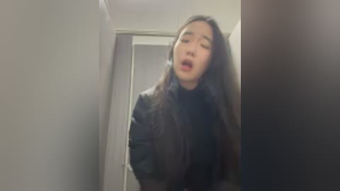 A blurry video of a young woman with long, dark hair, wearing a black jacket, standing in a narrow hallway with white walls and a partially open door behind her.