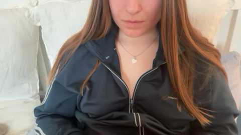 Video of a young woman with long, straight auburn hair, wearing a black zip-up jacket with a visible logo, seated in a softly lit bedroom with white bedding and a decorative pillow.