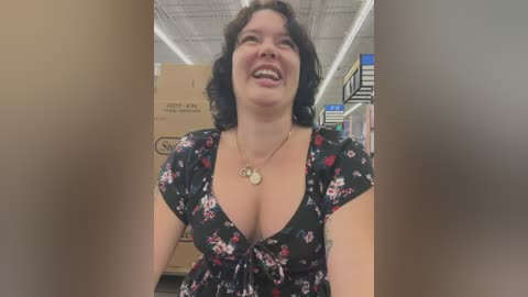 Video of a smiling, fair-skinned woman with short, dark hair, wearing a low-cut black floral dress. She stands in a brightly lit store aisle with a cardboard box and shelves in the background.